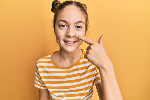 Красива брюнетка маленька дівчинка в повсякденній смугастій сорочці вказує пальцем на обличчя і ніс, усміхаючись весела. концепція краси
  - Фото, зображення