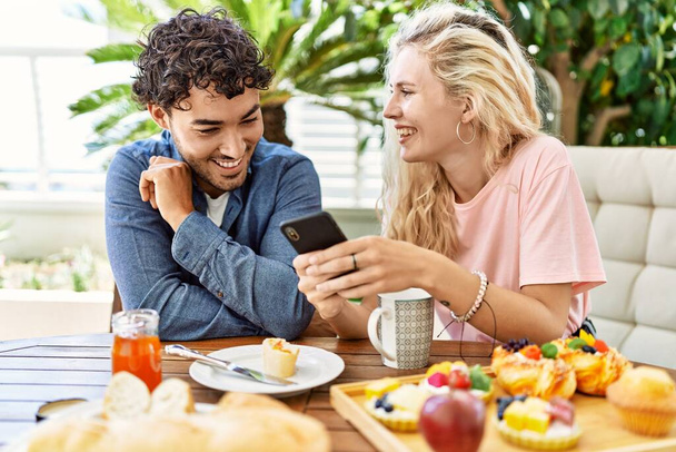 Giovane coppia che fa colazione e utilizza smartphone seduto sul tavolo in terrazza. - Foto, immagini