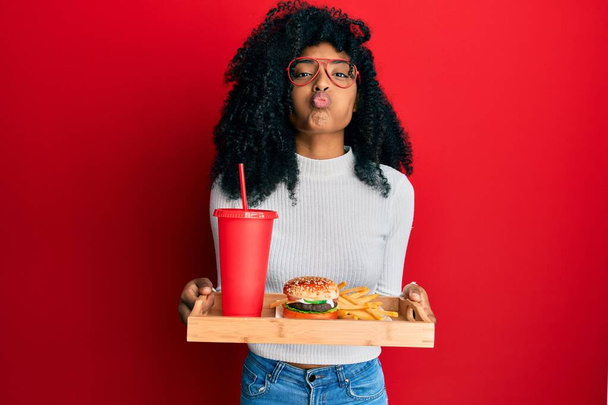 Afroamerikanerin mit Afrohaaren, die einen leckeren klassischen Burger mit Pommes frites und Limo mit lustigem Gesicht isst. Mund mit Luft aufgeblasen, Luft einfangen.  - Foto, Bild