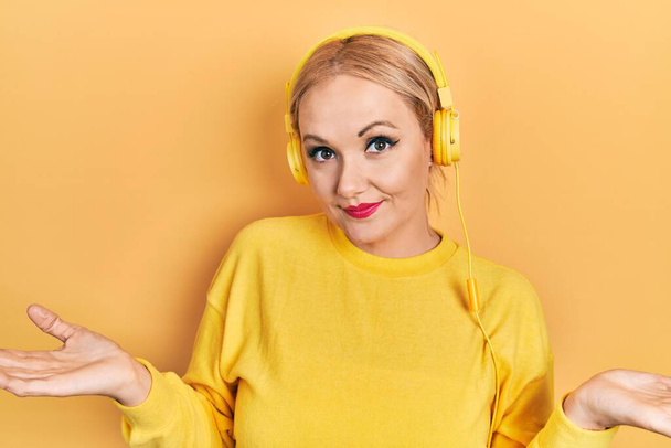 Молода блондинка слухає музику, використовуючи навушники безглузді і заплутані вирази з піднятими руками і руками. концепція сумніву
.  - Фото, зображення