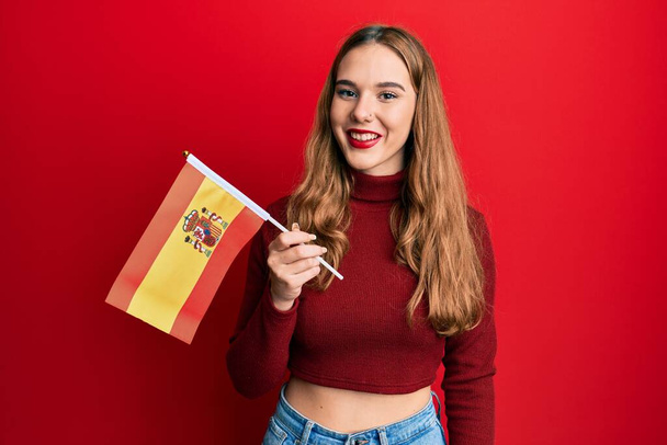 Mladá blondýna žena držící španělskou vlajku vypadá pozitivně a šťastně stojící a usmívající se s sebevědomým úsměvem ukazující zuby  - Fotografie, Obrázek