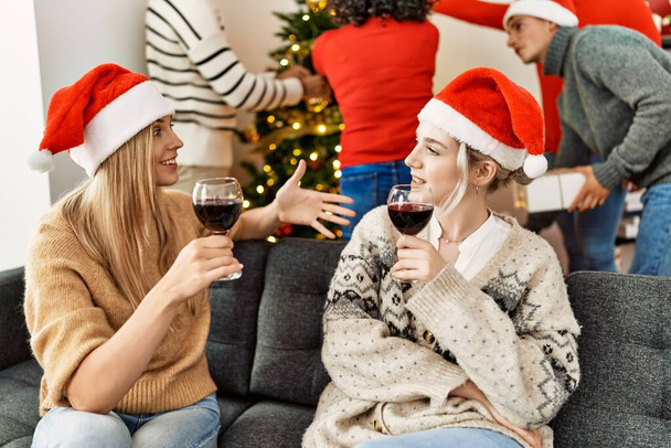 クリスマスを祝う若者のグループ。2人の女性が話して家でワインのグラスで乾杯. - 写真・画像