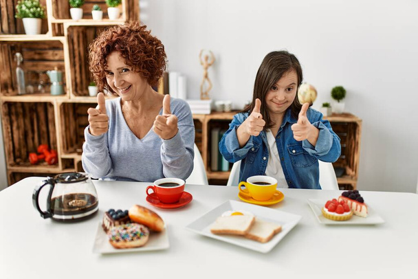 Familie van moeder en beneden syndroom dochter zitten thuis ontbijten wijzende vingers naar camera met gelukkig en grappig gezicht. goede energie en vibes.  - Foto, afbeelding
