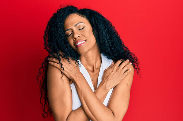 Middle age african american woman wearing casual white t shirt hugging oneself happy and positive, smiling confident. self love and self care  - Photo, Image