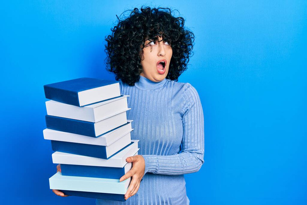 Young middle east woman holding a pile of books angry and mad screaming frustrated and furious, shouting with anger. rage and aggressive concept.  - 写真・画像