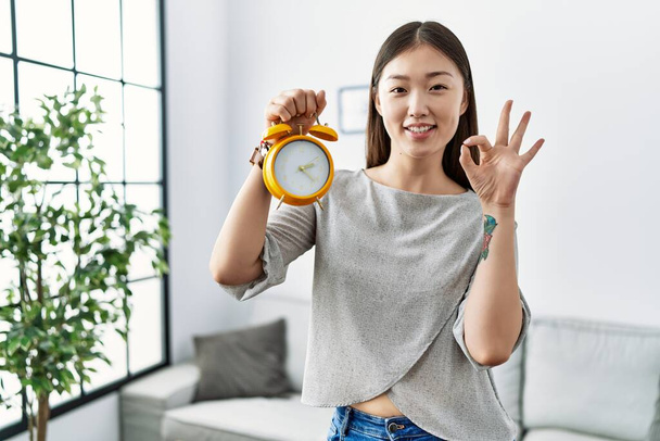 Young asian woman holding alarm clock doing ok sign with fingers, smiling friendly gesturing excellent symbol  - Valokuva, kuva
