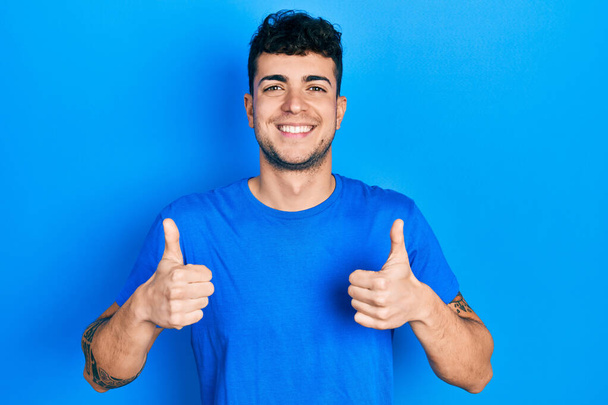 Jeune homme hispanique portant casual bleu t-shirt signe de succès faire un geste positif avec la main, pouces levés souriant et heureux. expression joyeuse et geste gagnant.  - Photo, image