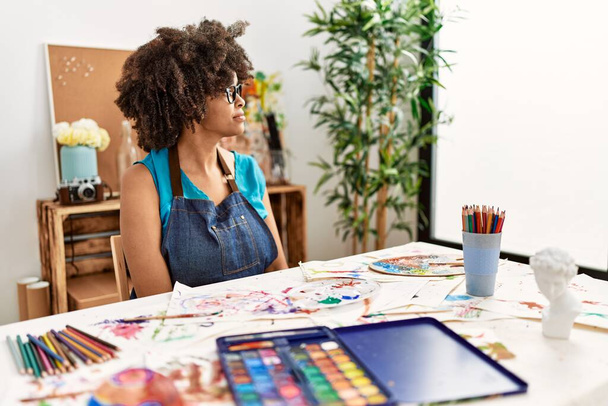 Beautiful african american woman with afro hair painting at art studio looking to side, relax profile pose with natural face with confident smile.  - Foto, afbeelding