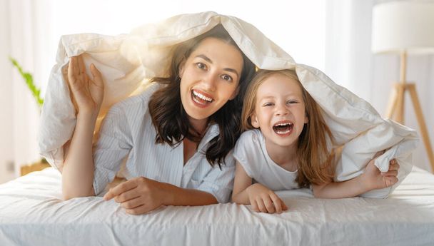 Ein nettes Mädchen und ihre Mutter genießen den sonnigen Morgen. Gute Zeit zu Hause. Kind wacht aus dem Schlaf auf. Familie spielt auf dem Bett im Schlafzimmer.  - Foto, Bild