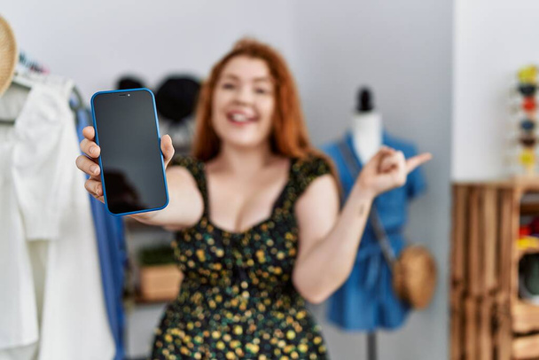 Giovane donna rossa al negozio al dettaglio che mostra lo schermo dello smartphone sorridente felice indicando con mano e dito di lato  - Foto, immagini