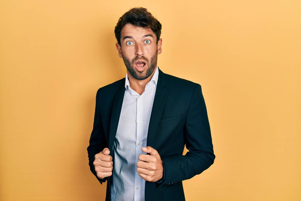 Handsome man with beard wearing business suit holding jacket in shock face, looking skeptical and sarcastic, surprised with open mouth  - Foto, afbeelding