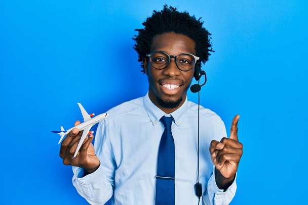 Jonge Afro-Amerikaanse reisagent met vliegtuig glimlachend met een idee of vraag wijzend met een vrolijk gezicht, nummer één  - Foto, afbeelding