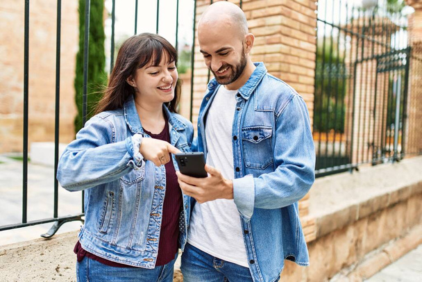 Νεαρό ζευγάρι ισπανόφωνων χαμογελά χαρούμενος χρησιμοποιώντας smartphone στην πόλη. - Φωτογραφία, εικόνα