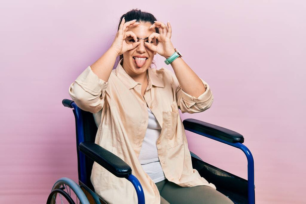 Young hispanic woman sitting on wheelchair doing ok gesture like binoculars sticking tongue out, eyes looking through fingers. crazy expression.  - Zdjęcie, obraz