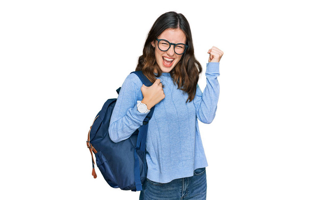 Молодая красивая девушка носит студенческий рюкзак кричать гордо, празднуя победу и успех очень взволнован с поднятыми руками  - Фото, изображение