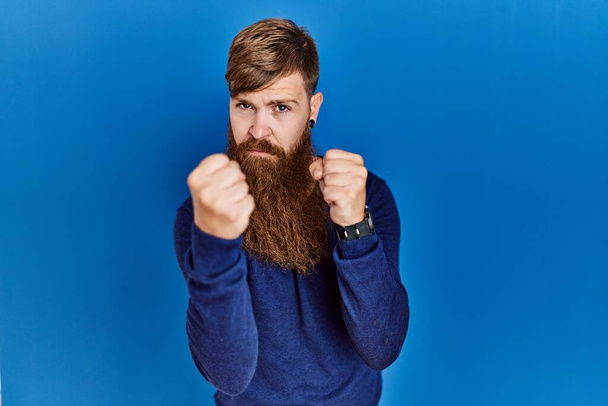 Рыженький с длинной бородой, в обычном голубом свитере на синем фоне, готовый драться жестом защиты кулака, сердитый и расстроенный, боится проблем  - Фото, изображение