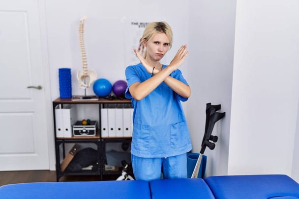 Mujer fisioterapeuta joven que trabaja en la clínica de recuperación del dolor expresión de rechazo cruzando los brazos haciendo signo negativo, cara enojada  - Foto, imagen