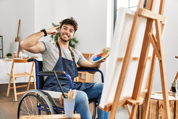 Joven hombre hispano sentado en la pintura en silla de ruedas en el estudio de arte sonriendo señalando a la cabeza con un dedo, gran idea o pensamiento, buena memoria  - Foto, Imagen