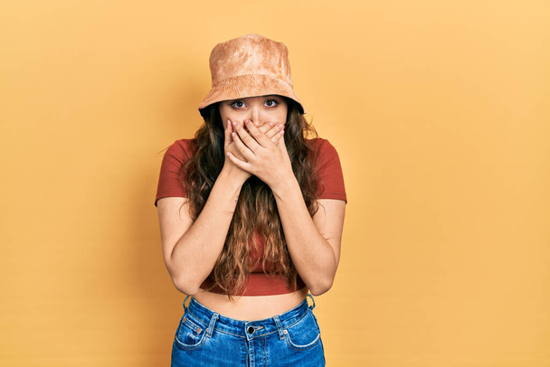 Young hispanic girl wearing casual clothes and hat shocked covering mouth with hands for mistake. secret concept.  - Photo, Image