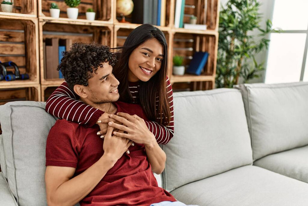 Junges lateinisches Paar lächelt glücklich und umarmt sich zu Hause auf dem Sofa. - Foto, Bild