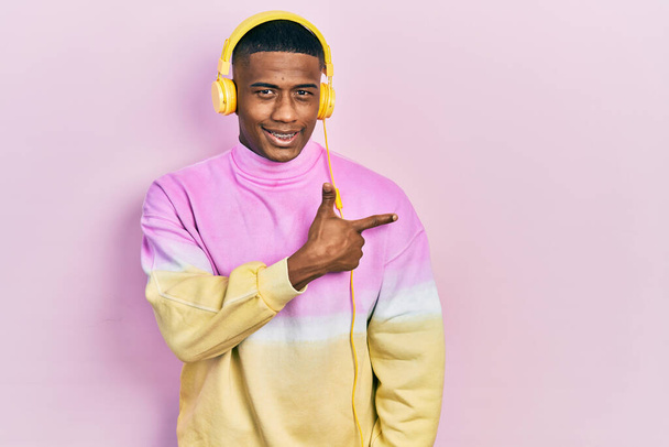 Молодий чорний чоловік слухає музику в навушниках веселий з посмішкою обличчя, спрямованою рукою і пальцем в сторону з щасливим і природним виразом на обличчі
  - Фото, зображення