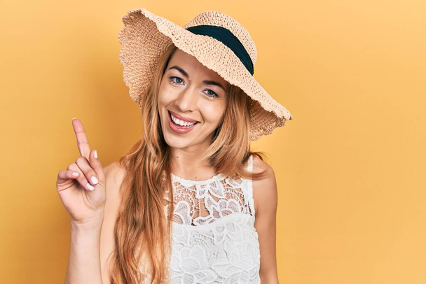 Młoda kaukaska kobieta w letniej czapce pokazuje i wskazuje palcem numer jeden, uśmiechając się pewny siebie i szczęśliwy.  - Zdjęcie, obraz