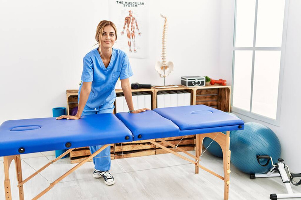 Junger kaukasischer Physiotherapeut lächelt glücklich auf Tisch in der Klinik gelehnt - Foto, Bild
