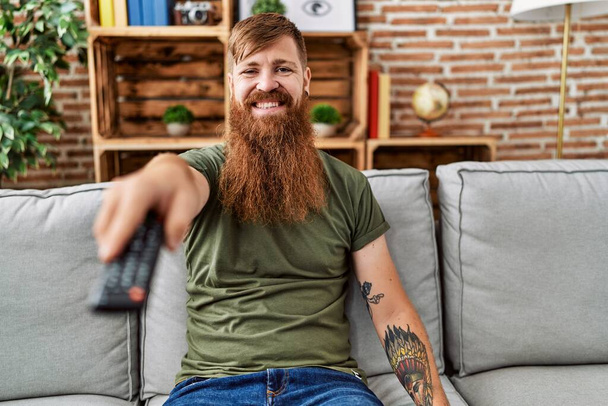 Vörös hajú férfi hosszú szakállal kezében televízió távirányító néz pozitív és boldog álló és mosolygó magabiztos mosollyal mutatja fogak  - Fotó, kép
