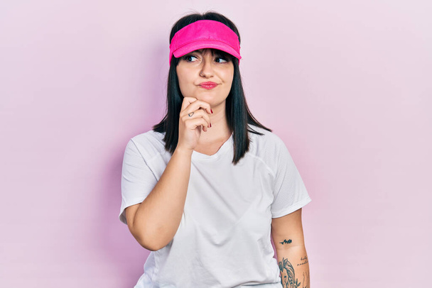 Young hispanic woman wearing sportswear and sun visor cap serious face thinking about question with hand on chin, thoughtful about confusing idea  - Photo, Image