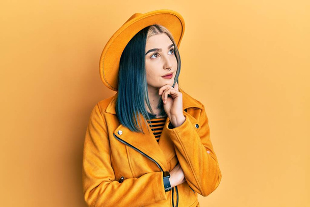 Young modern girl wearing yellow hat and leather jacket with hand on chin thinking about question, pensive expression. smiling and thoughtful face. doubt concept.  - Photo, Image