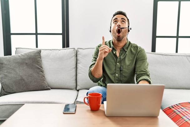 Young hispanic man with beard wearing call center agent headset working from home amazed and surprised looking up and pointing with fingers and raised arms.  - Photo, Image