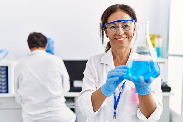 Middelbare leeftijd man en vrouw partners dragen wetenschapper uniform houden reageerbuis in laboratorium - Foto, afbeelding