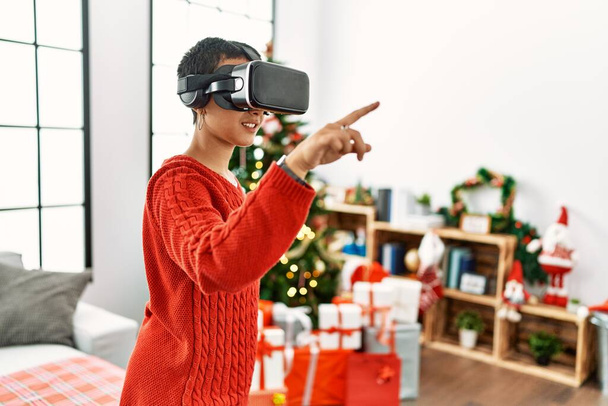 Young hispanic woman playing video game using vr glasses standing by christmas tree at home - Photo, image