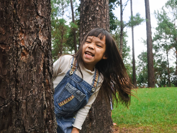 かわいい女の子は大きな木の後ろに立って、カメラで笑顔。屋外で遊んでいる幸せな子供。一緒に過ごした家族. - 写真・画像