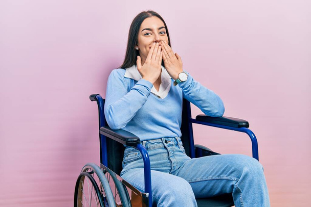 Schöne Frau mit blauen Augen, die im Rollstuhl sitzt, lacht und verlegen kichert, den Mund mit Händen bedeckt, Klatsch und Skandalkonzept  - Foto, Bild