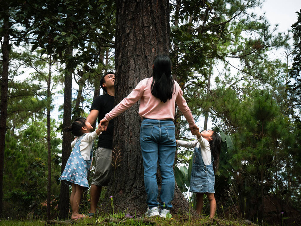 Ευτυχισμένες οικογένειες στέκονται κρατώντας ο ένας το χέρι του άλλου γύρω από ένα μεγάλο δέντρο. Ευτυχισμένη οικογένεια που παίζει έξω. Έννοια διατήρησης του περιβάλλοντος. - Φωτογραφία, εικόνα