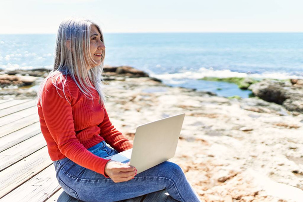 Μέση ηλικία γκρίζα μαλλιά γυναίκα χρησιμοποιώντας φορητό υπολογιστή κάθεται στον πάγκο στην παραλία. - Φωτογραφία, εικόνα