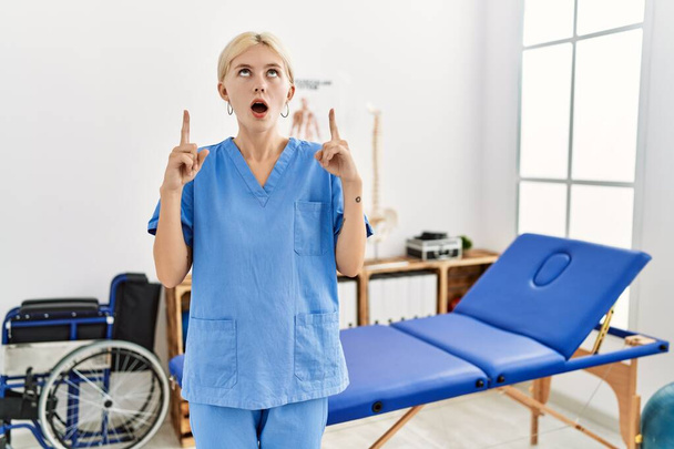 Hermosa fisioterapeuta caucásica mujer que trabaja en la clínica de recuperación del dolor sorprendido y sorprendido mirando hacia arriba y señalando con los dedos y los brazos elevados.  - Foto, imagen