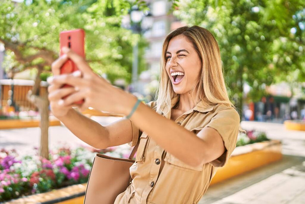 Young blonde girl smiling happy making selfie by the smartphone at the city. - Фото, изображение