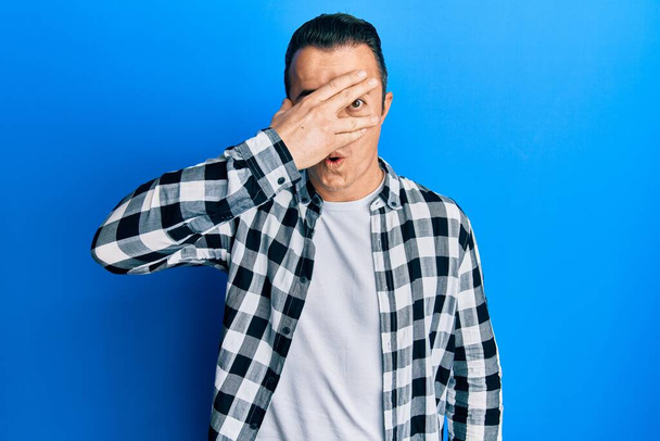 Handsome young man wearing casual shirt peeking in shock covering face and eyes with hand, looking through fingers afraid  - Photo, image