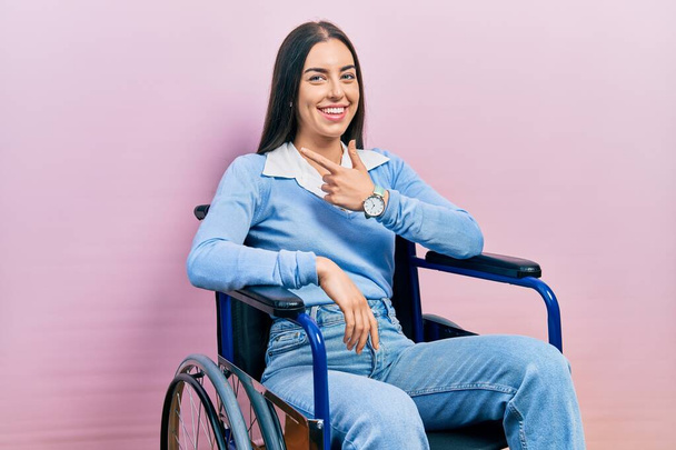 Mulher bonita com olhos azuis sentada em cadeira de rodas alegre com um sorriso no rosto apontando com a mão e o dedo para o lado com expressão feliz e natural  - Foto, Imagem