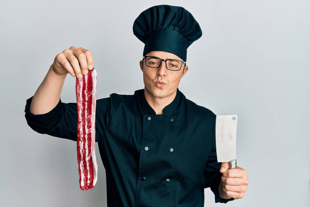 Handsome young man wearing chef uniform holding bacon and knife looking at the camera blowing a kiss being lovely and sexy. love expression.  - Fotografie, Obrázek