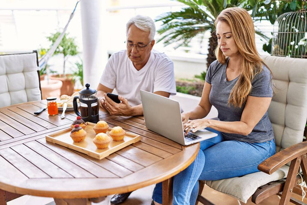Keski-ikäinen latinopari aamiaisella älypuhelimella ja kannettavalla tietokoneella terassilla. - Valokuva, kuva