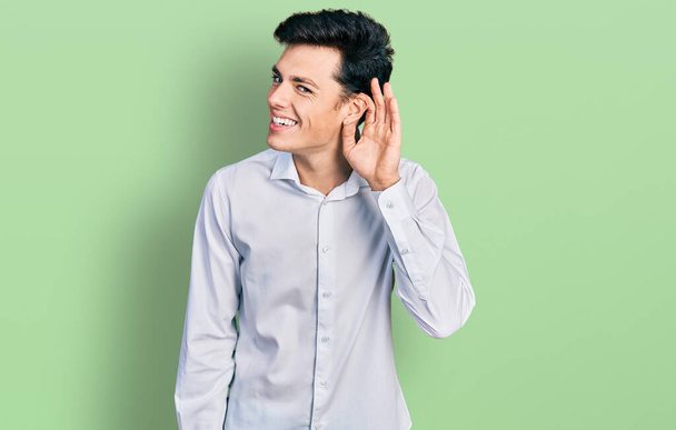 Jovem homem de negócios hispânico vestindo roupas de negócios sorrindo com a mão sobre o ouvido ouvindo um rumor ou fofocas. conceito de surdez.  - Foto, Imagem