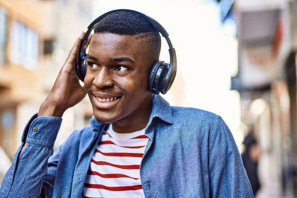 Νεαρός Αφροαμερικάνος χαμογελώντας χαρούμενος χρησιμοποιώντας ακουστικά στην πόλη. - Φωτογραφία, εικόνα