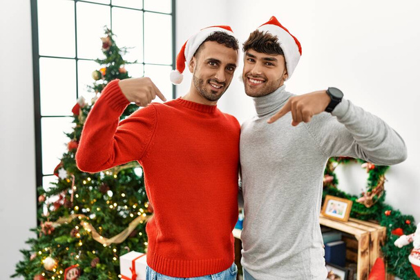 Young gay couple standing by christmas tree wearing hat looking confident with smile on face, pointing oneself with fingers proud and happy.  - Photo, Image
