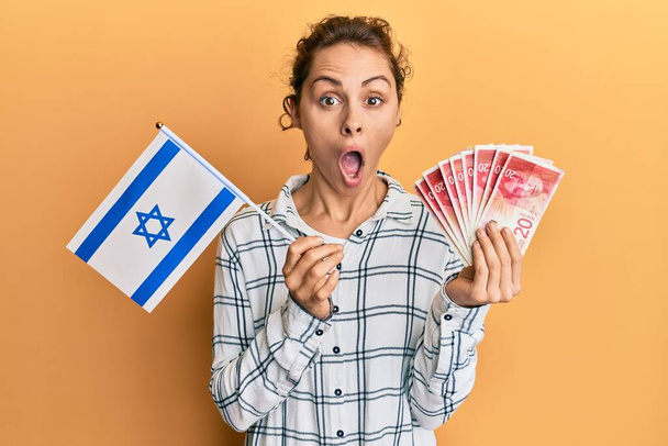 Młoda brunetka trzymająca flagę Izraela i sykle banknotów przerażona i zszokowana zaskakującą ekspresją, strachem i podnieconą twarzą.  - Zdjęcie, obraz