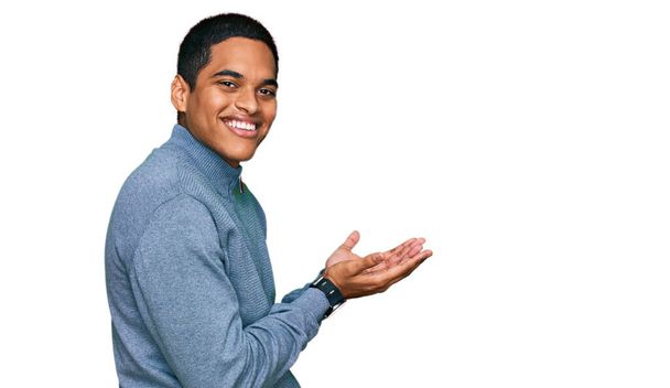 Mladý pohledný hispánec nosí ležérní mikinu ukazující stranou s rukama otevřenýma dlaněmi ukazující kopírovací prostor, prezentující reklamu s nadšeným úsměvem  - Fotografie, Obrázek
