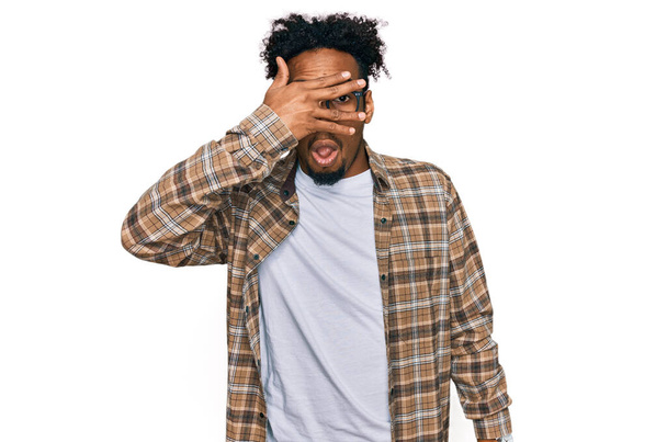 Young african american man with beard wearing casual clothes and glasses peeking in shock covering face and eyes with hand, looking through fingers with embarrassed expression.  - Fotó, kép