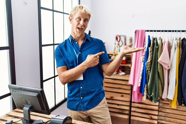 Молодий блондин, який працює менеджером в роздрібному магазині, здивований і посміхається камері, представляючи рукою і вказуючи пальцем
.  - Фото, зображення